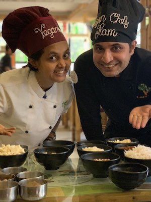 Chef Susmitha and Chef Ram smiling at the camera and bending over and pointing to ingredients.