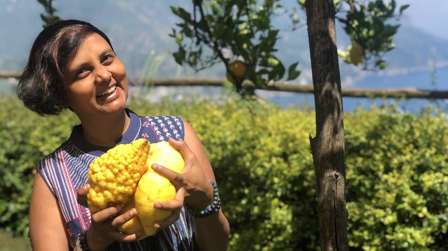 Short haired Indian girl holding large Italian lemons and smiling at the camera. Head tilted slightly. Blurred trees and plants in the background.