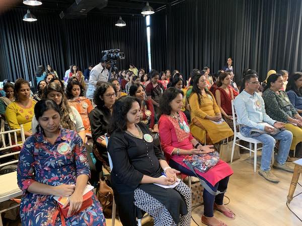 Lots of women, with a couple of men, sitting on chairs, eyes closed, meditating together.