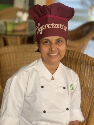 Susmitha, sitting, facing camera. Smiling wide. Wearing a Chef Coat. Chef Cap with the caption Veganosaurus stitched on it.