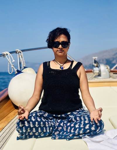 Susmitha sitting cross legged on a boat. Palms on knees, facing upwards. Index and thumbs touching in gyaana mujra. Short hair. Wearing sunglasses.