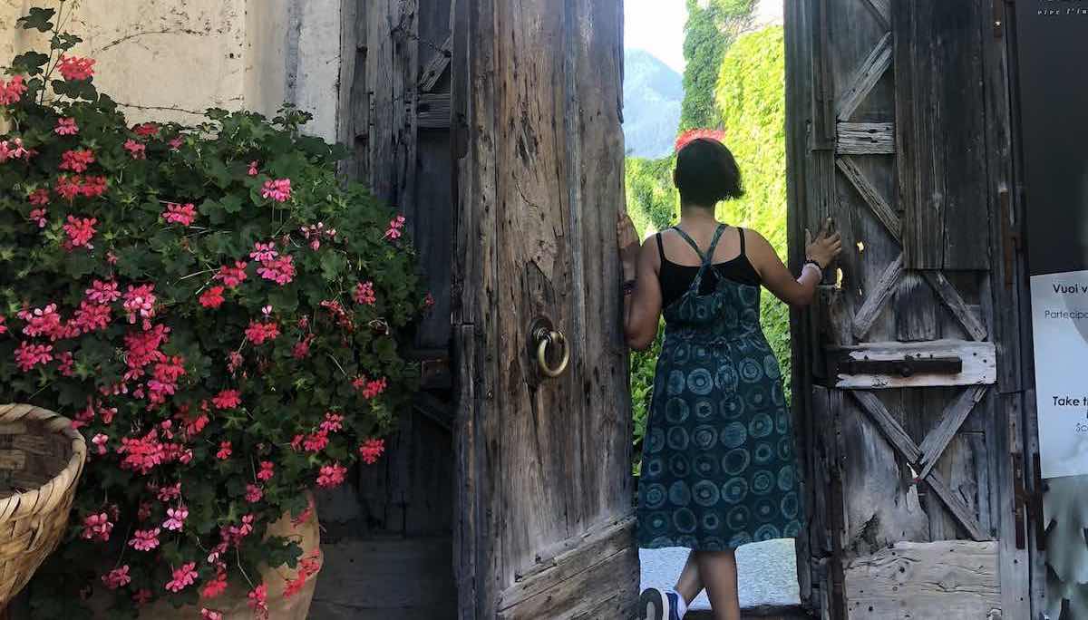 Girl in a dress, standing at a wooden doorway, back to the camera. Large flower pot on the left. Greenery in the distance outside the door.