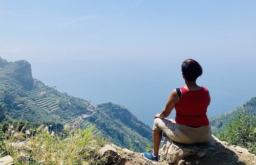 Mountaintop scenery. Wide skies and hills far away. At the bottom right corner of the photo, there's a girl sitting on a rock with her back to the camera.