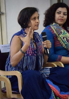 Susmitha Subbaraju sitting in a chair and speaking. Mic in one hand, other palm raised up and turned towards her in communication. Fellow panelist lady sitting next to her and looking at her.