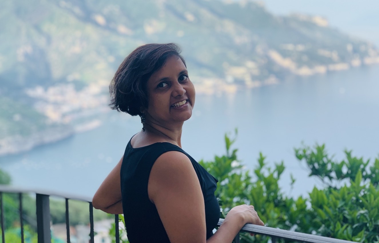 Susmitha, holding railings at a cliffside, looking over her shoulder at the camera. Water and mountain scenery in the background.