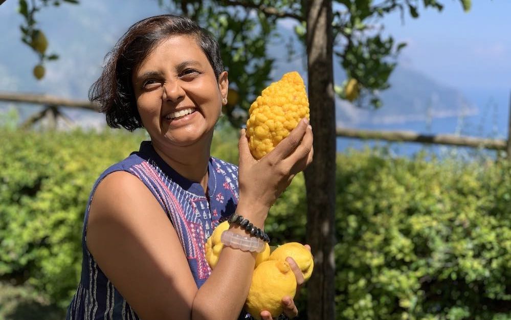 Susmitha holding large lemons and smiling at the camera. Greenery in the background. Location cliffside villa at Ravello, Italy.