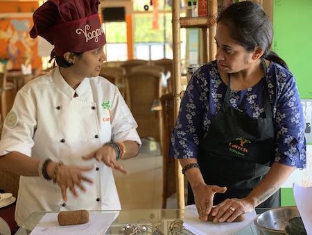Indian vegan woman chef teaching cooking to another Indian lady. Both standing on one side of a table, heads turned towards each other.