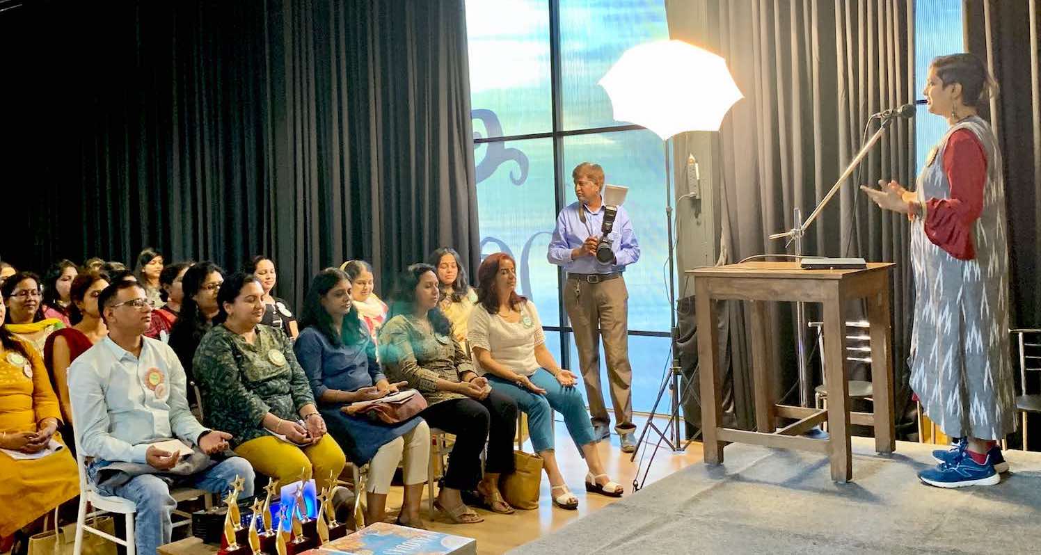 Wide view of Susmitha Subbaraju at OMBC Discover. Speaking into a standing mic on stage. Audience in chairs, about to start meditating.