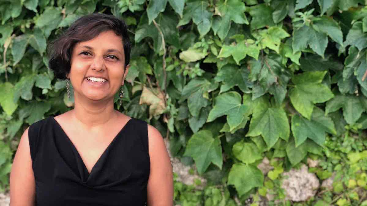 Susmitha Veganosaurus, vegan business coach, in a black dress, short hairstyle. Smiling at the camera. Standing in front of a wall of green vines and leaves.