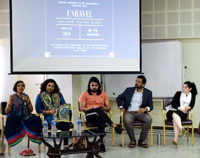 Susmitha, seated crosslegged on a chair, holding a mic and speaking. Four other panelists also seated on chairs next to her on stage.