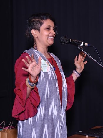 Susmitha standing and speaking into a stand mic. Arms spread out. Smiling. Wearing long, frilled sleeves and crystal bracelets. Super short hairstyle.