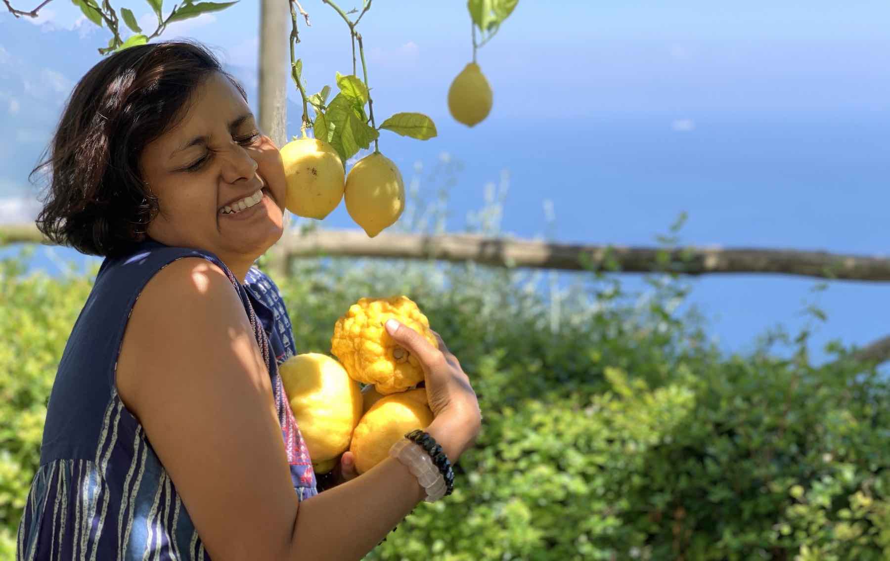 Susmitha, short haired Indian lady. Holding lemons tightly in a hug and pressing cheek to lemons on a tree with eyes closed and smiling happily. Greenery, water and blue skies in the background.