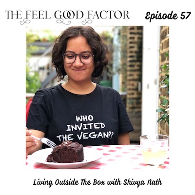 The Feel Good Factor, episode 57. Digital nomad and travel writer Shivya Nath, wearing glasses and a "WHO INVITED THE VEGAN?" tee. Seated at an outdoor table, looking down with a smile and reaching with a fork to a chocolate cake. Living Outside The Box with Shivya Nath.