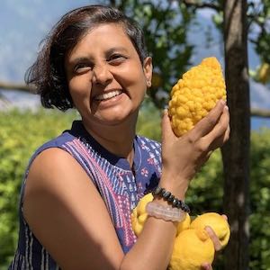 Susmitha holding large lemons and smiling at the camera. Greenery in the background. Location cliffside villa at Ravello, Italy. Veganosaurus Newsletter.