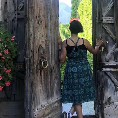 Girl in a dress, standing at a wooden doorway, back to the camera. Large flower pot on the left. Greenery in the distance outside the door.
