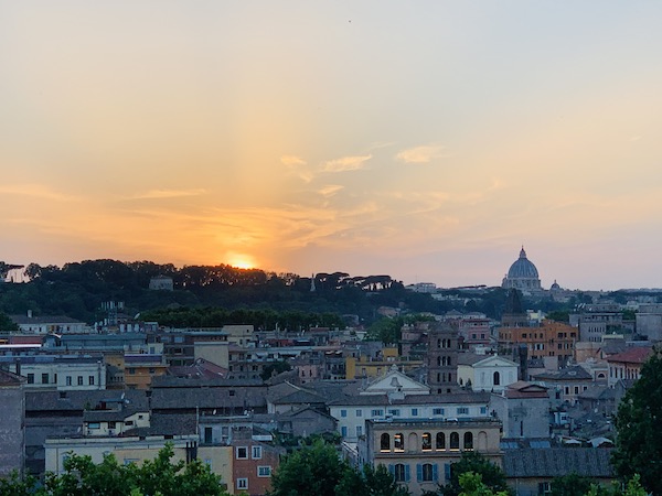 View of Rome, Italy from the Giardino Degli Aranci. Top half: the sun setting in the sky. Bottom half: lots of old buildings and some trees.