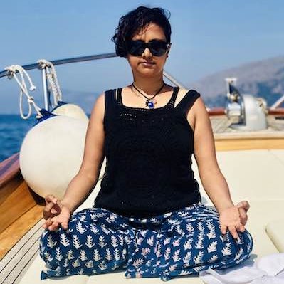Susmitha sitting cross legged on a boat. Palms on knees, facing upwards. Index and thumbs touching in gyaana mujra. Short hair. Wearing sunglasses.