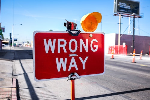 Large, red street sign that says "WRONG WAY". Road in the background with traffic cones, lights, hoarding.