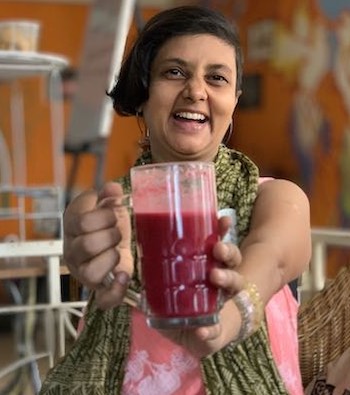 Vegan influencer Susmitha Veganosaurus holding out a glass of what appears to be beet juice towards the camera and laughing. Blurred background.