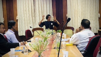 Chef Susmitha Veganosaurus conducting a vegan consultation session. Hands raised as she speaks. Men sitting at a conference table and listening intently, learning how to make vegan food.