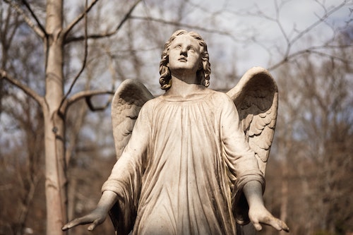 Stone statue of an angel. Looking up. Sunrays falling on her. Blurred background with tress (no leaves on them). Surrender to a higher power.