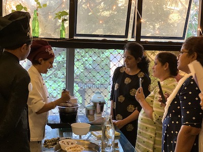 Learn how to make vegan food, cooking demonstration. Left: Chef Susmitha in white chef coat cooking, Chef Ram in black chef coat watching. Centre: Glass table with ingredients, blender, induction stove. Right: Students, three women, watching and taking photos on their phones.