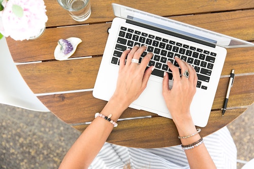 top view, laptop on round wooden table. Woman's hands with rings and bracelets shown typing on it. Pen, amethyst crystal, glass of water, flowers in a vase on the table around the laptop. Productivity.