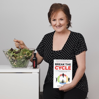 Karen Warwick, short haired woman, seated and holding her book Break The Cycle. Large bowl of green salad next to her, she's holding up a forkful and smiling at the camera.