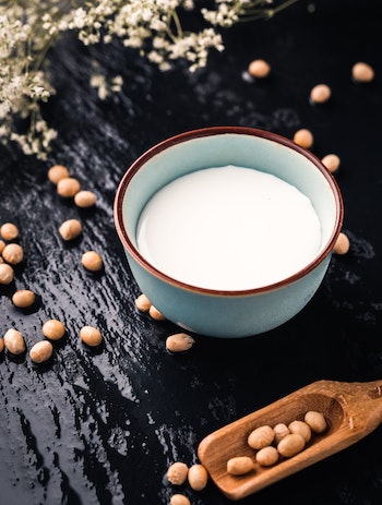 Black, textured stone background. Ceramic bowl with peanut yogurt in it. Peanuts strewn around it. Elongated wooden scoop with peanuts in it lying next to the bowl of yogurt. Vegan dairy alternatives.