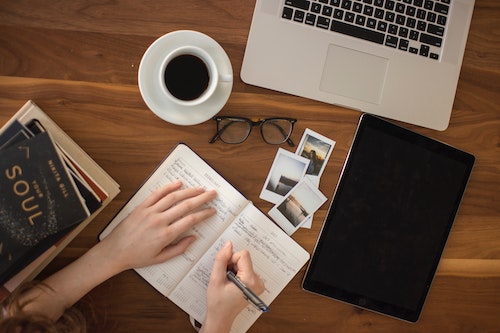 Top view of deck with laptop, black coffee in a cup, books on the side, black and white photos, planner, glasses, and a person's hands writting in a journal. Business Coaching.