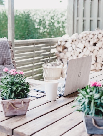 Outside work table. Angled close up view. Flowering plants and laptop on a wooden desk. Beverage in a mug on the side. Vegan business coaching.