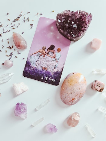 White background with purple tarot card, 3 of cups, showing an illustration of three women from behind. Various crystals in shades of purples, lavenders, and pinks surround the card.