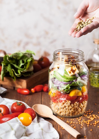 Buddha bowl glass jar with layers of grains, veggies, seeds, and tofu on a wooden table. Tomatoes and greens around it. wooden spoon on the table. Top right corner there's a hand pouring seeds into the glass jar. Vegan cooking consultation.
