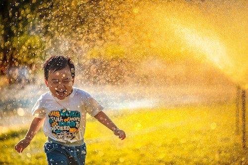 Little kid laughing freely and running in water spray. The water's reflecting the sunlight in a golden yellow hue. Authentically joyful energy in the picture.