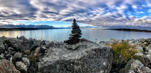 Gray stones piled up on a rock with a beautiful blue lake in the background and a blue sky with white clouds. Surreal meditative feel to the image.