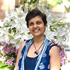 Shorth haired Indian lady, beaming a wide smile. Flowers in the background. Vegan business coach and chef Susmitha Veganosaurus