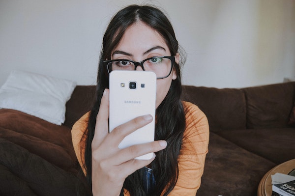 Woman sitting with hunched shoulders, wearing black glasses, holding her phone up to her face and staring at it with dull eyes. Stop phone addiction.