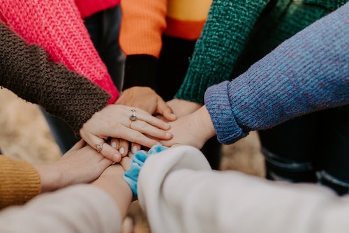Many hand coming together in the middle with palms placed on top of each other. All wearing colourful, long sleeved sweaters. The top hand has finger rings.