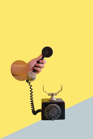 Yellow wall, hand coming out through a round hole and holding up the receiver of an antique rotary telephone