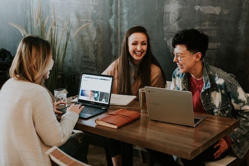 Three young people sitting around a table with laptops open in front of them. Talking and laughing together. Enjoying their work.