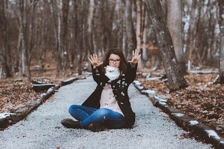 Caucasion woman with dark hair and glasses sitting on a path in a wooded area, facing thecamera with both arms lifted and palm facing out towards the camera.