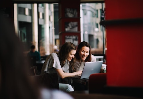 Two women seated at a table, working together on a laptop and laughing. Friendship and collaboration in business.