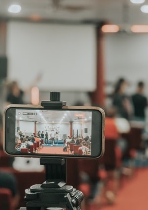 Mobile phone in landscape mode, shooting a video of a speaker on stage and the audience, blurred background showing people.