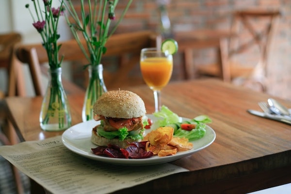 Vegan friendly restaurant table with burger, fries and salad on a plate, goblet of orange juice on the side. Vases and chairs in the background.