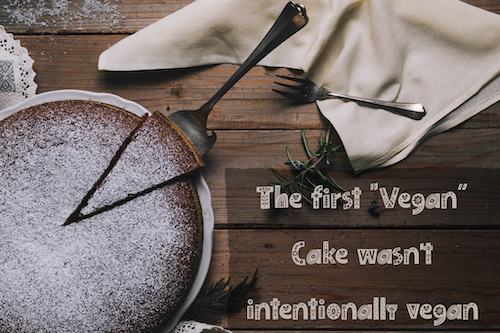 Chocolate cake on a plate with a slice  cut and slightly moved away from the main cake. Set on a slatted wooded table with napkins, forks and herbs strewn on it. Text: The first "Vegan" Cake wasn't intentionally vegan