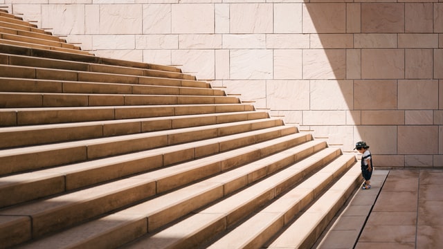 Toddle standing in front of a flight of large concrete stairs. Looking down at the first stair. It's a big challenge.