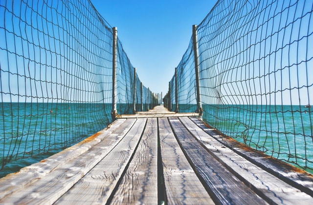 Brown wooden bridge with green safety net fence on either side, over turquoise blue water, blue sky in the horizon.