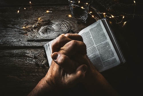 Hands clasped over a book on a dark table, in prayer. Representing the Sanskrit mantra of surrender, Idam na mama.