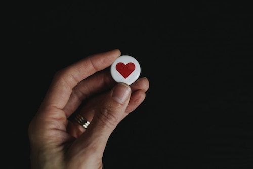 Close up of a hand holding a small white button with a red heart on it. Represents seeking validation and likes on social media.