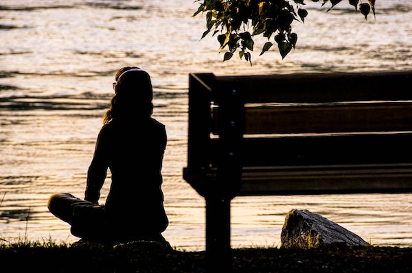 Silhouette of of woman sitting in front of a stream and meditating. Divine grounding meditation.